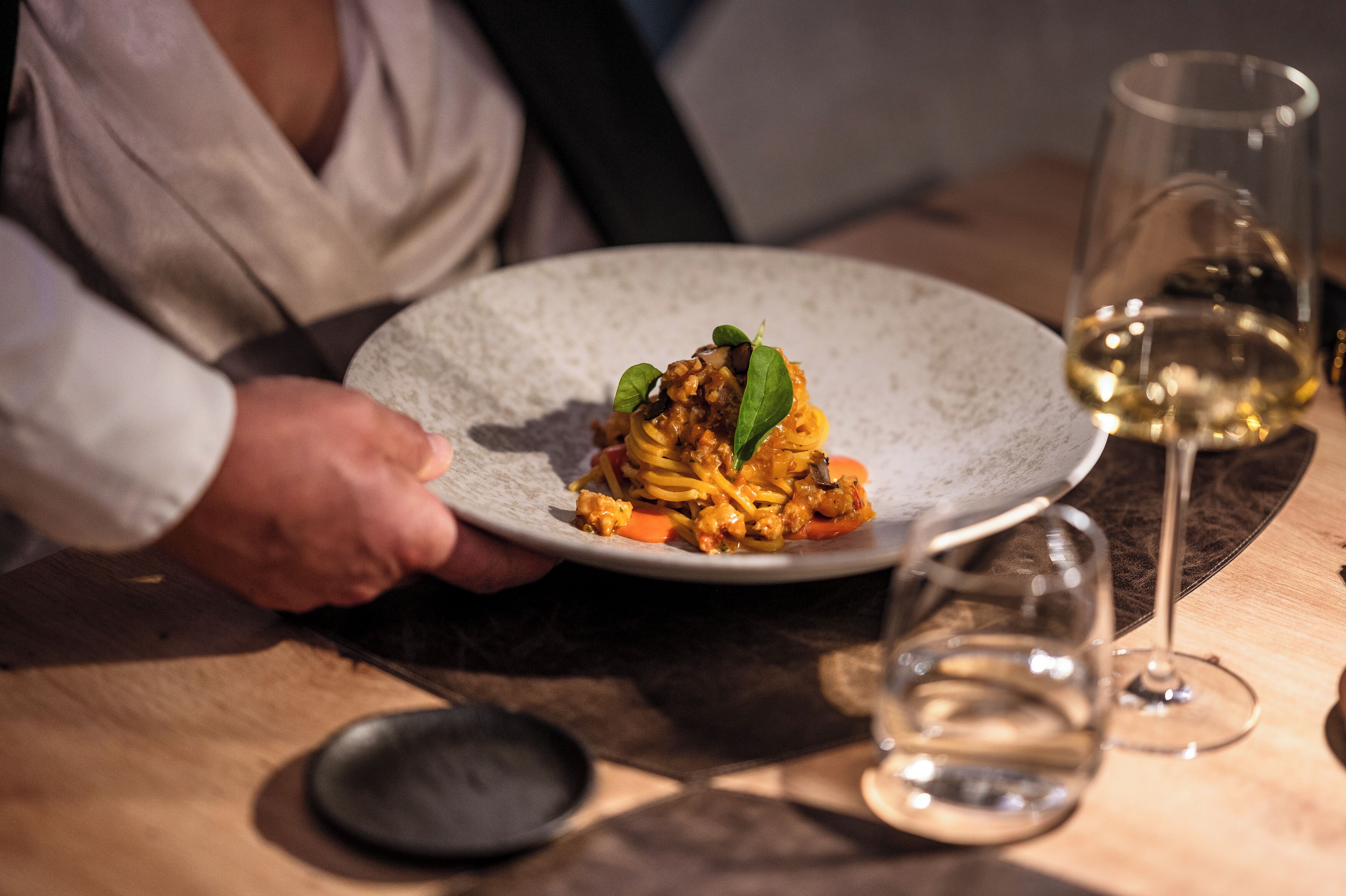 A person holding a plate of food at a table with a wine glass.