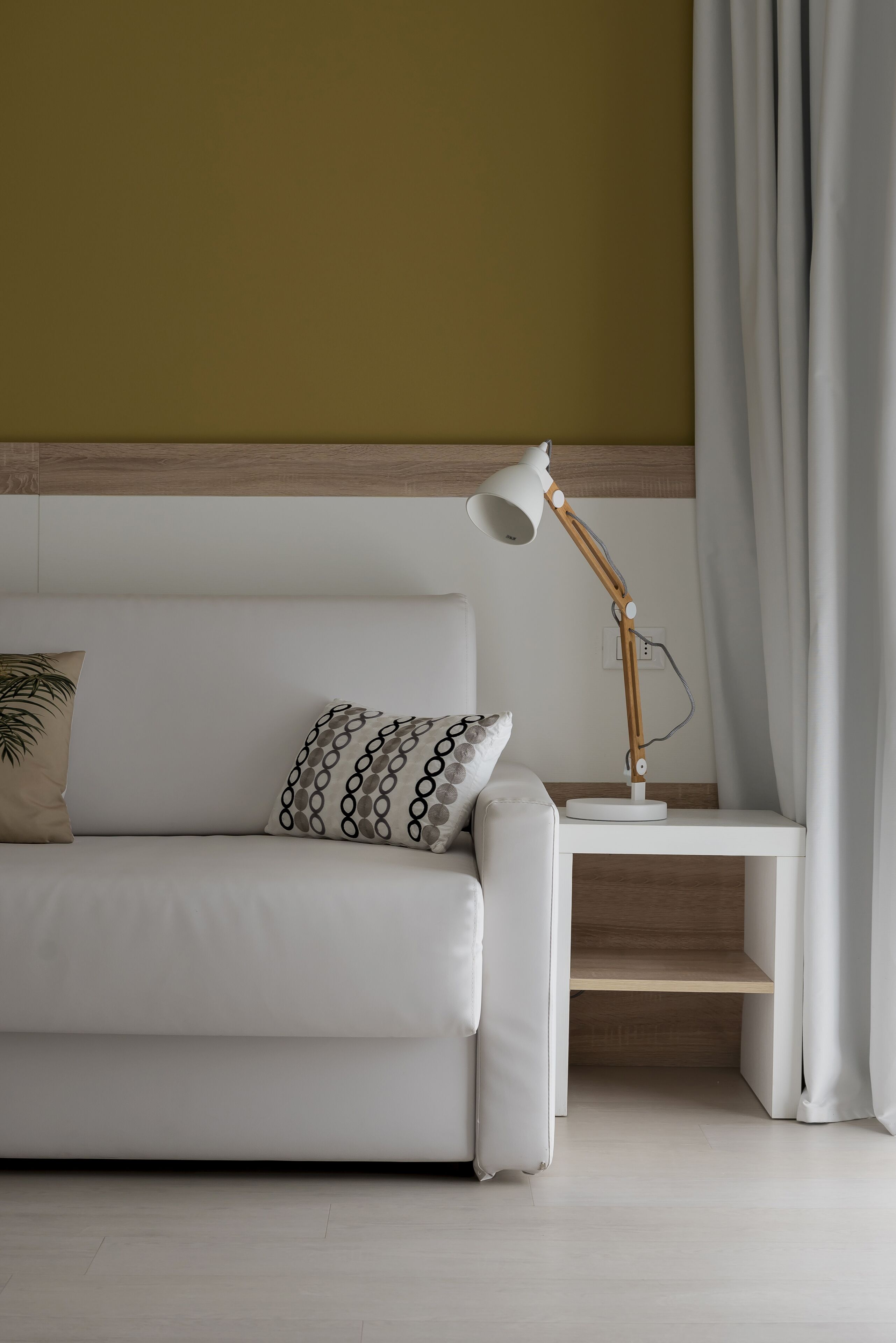 Un sofá blanco con almohadones estampados, una mesita de madera con una lámpara de escritorio y una pared lisa con un panel de madera.
