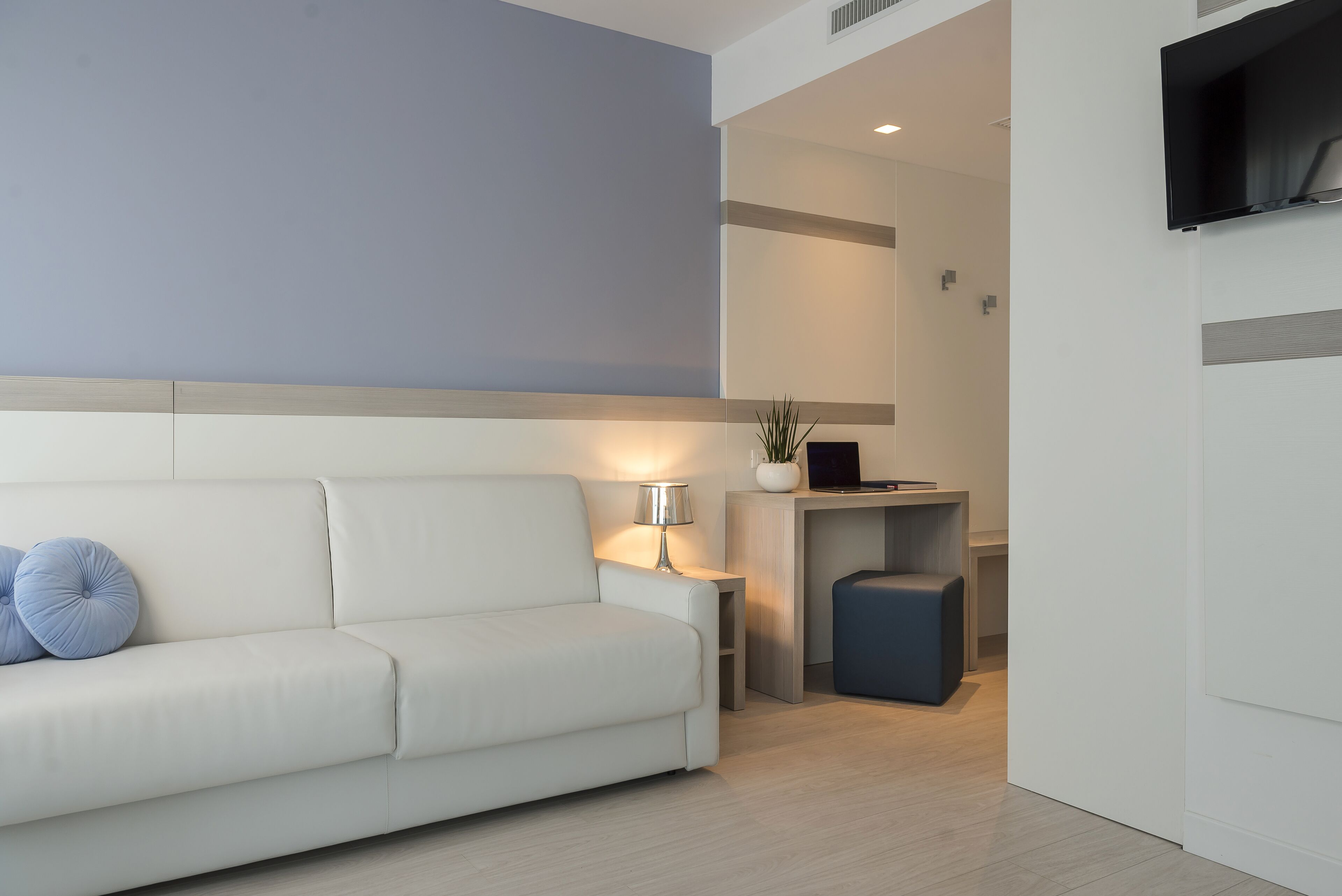 Una sala de estar moderna con un sofá blanco, una mesita de noche con una lámpara, y una pequeña zona de escritorio con un televisor y una planta en maceta.