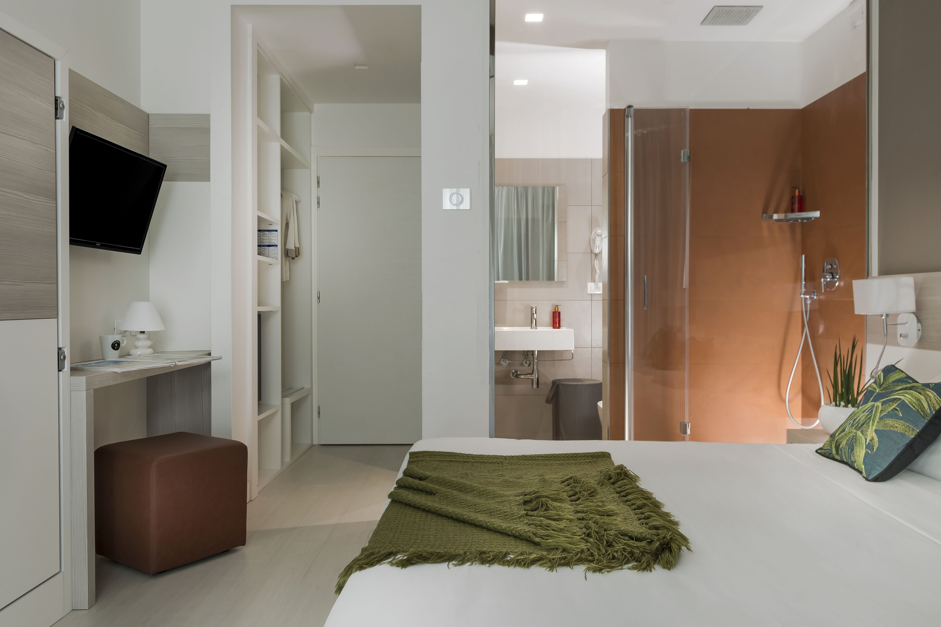 A modern hotel room with a bed, a desk, a TV, and a bathroom visible through an open door.