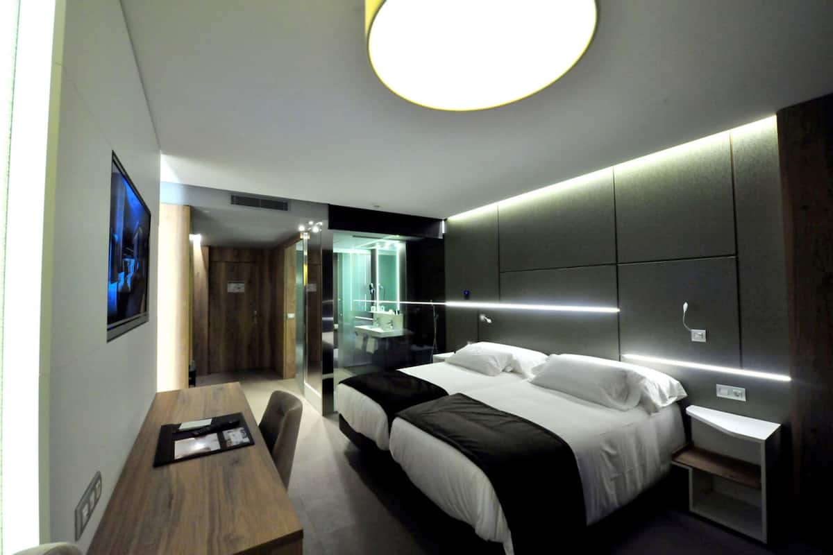 A modern hotel room with a large bed, bedside tables, a desk, and a glass partition separating the room from the bathroom.