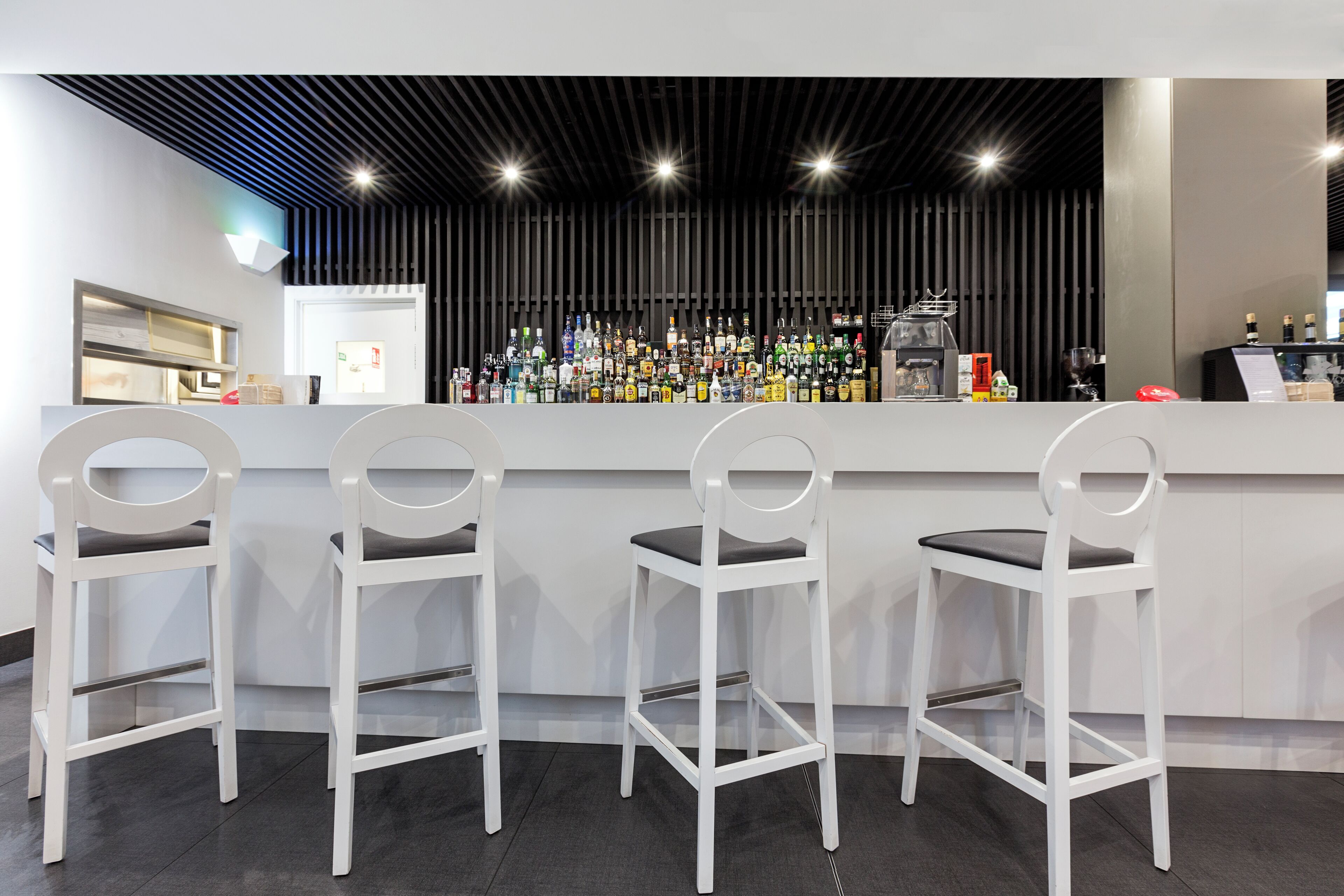 Eine Bar mit drei weißen Barhocker, einer Theke mit verschiedenen Flaschen und einem modernen Deckenstil.