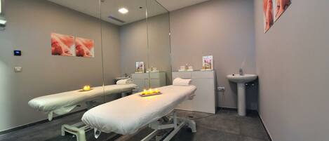 A clinical examination room with two examination tables, a large mirror, a sink, and a wall-mounted poster.