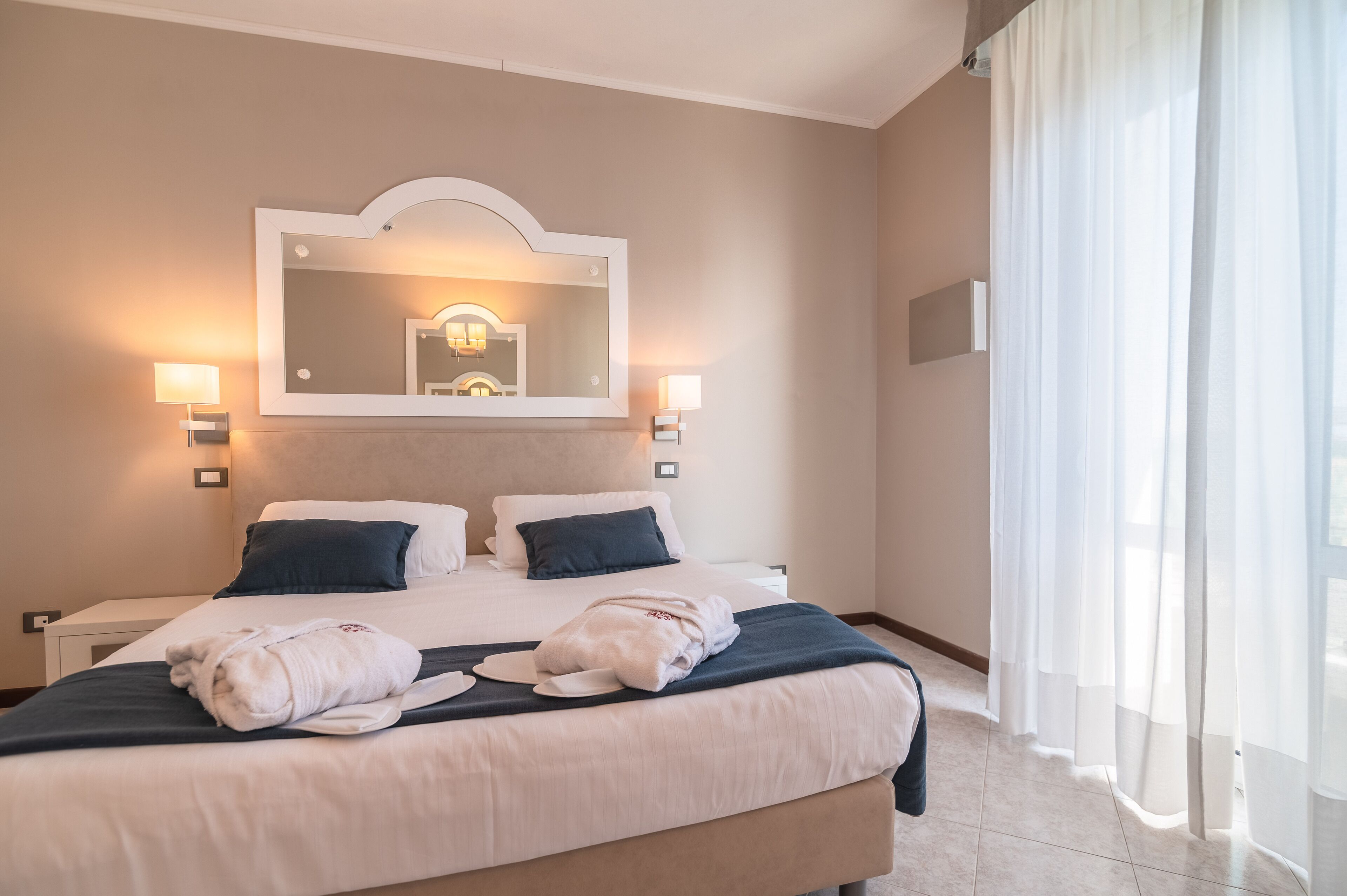 A hotel room with a large bed, two bedside lamps, a mirror above the bed, and sheer curtains on the window.