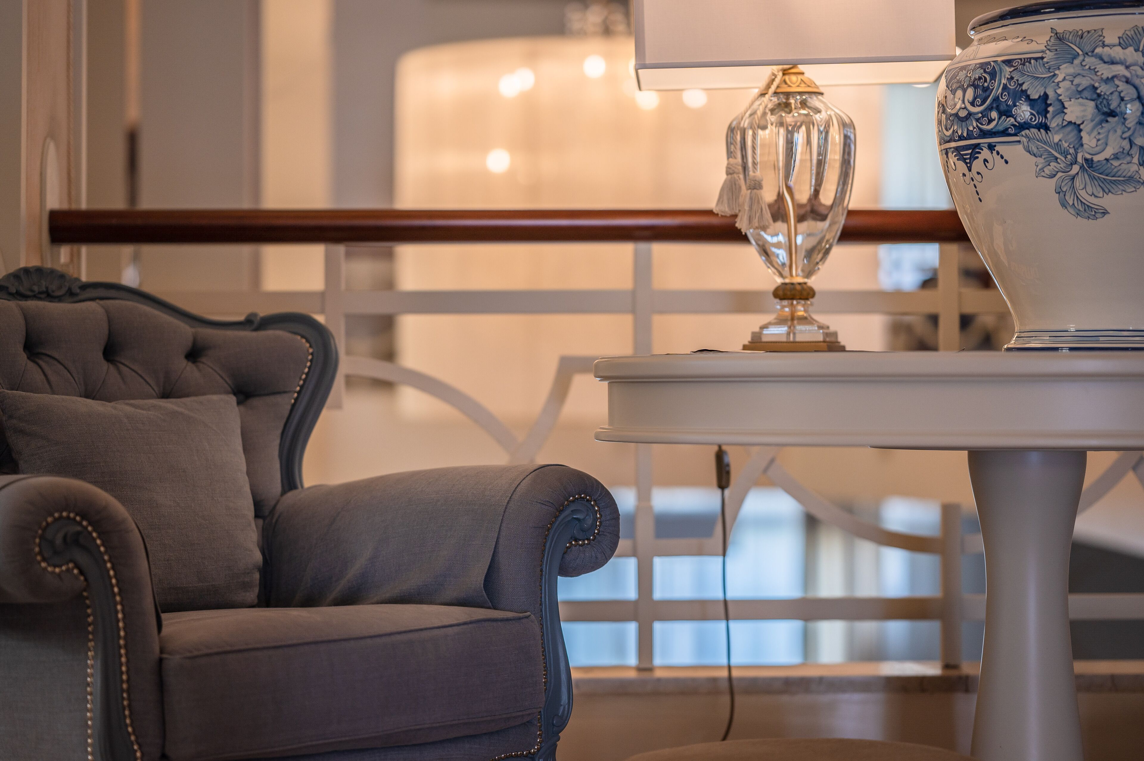 A room with a tufted armchair, a table with a decorative lamp, and a blue and white patterned vase.
