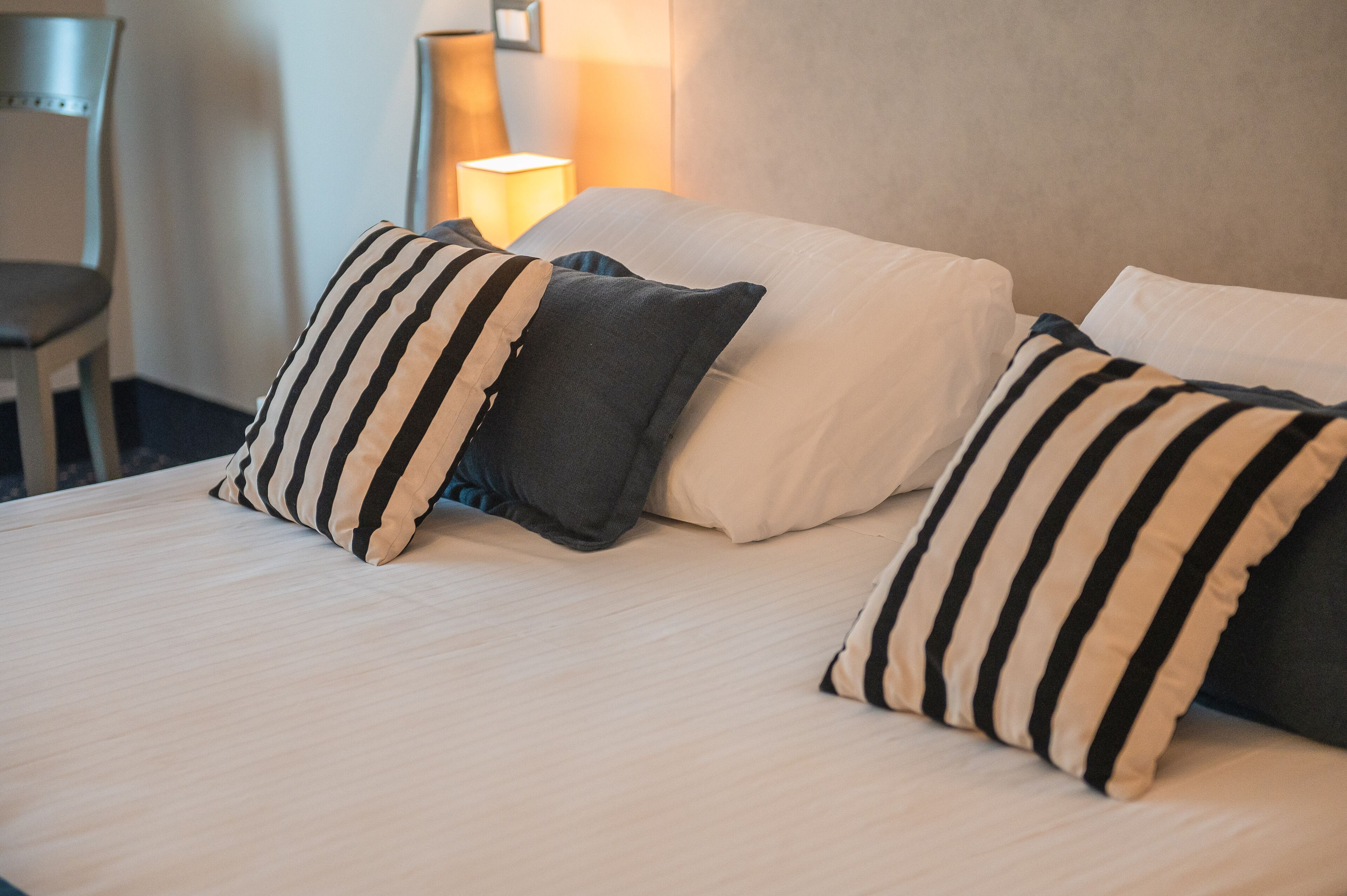 A neatly made bed with striped and solid pillows, a chair, and a bedside lamp.