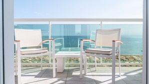 En balkon med to hvide stole og et lille bord, med udsigt over et klart, blåt hav og en klippefyldt kystlinje.