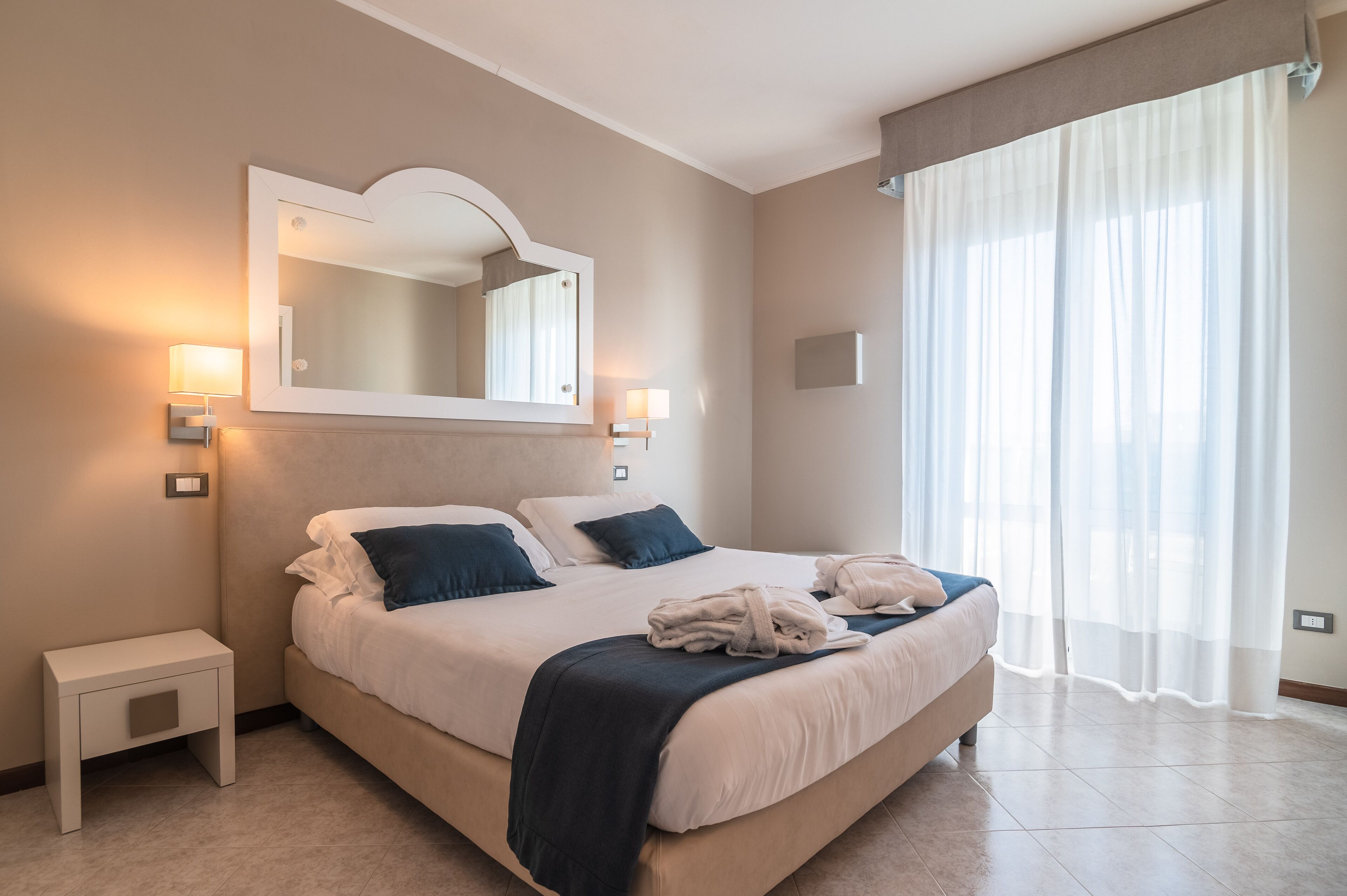 Habitación de hotel con una cama grande, lámparas de noche, un espejo y una ventana con cortinas translúcidas.