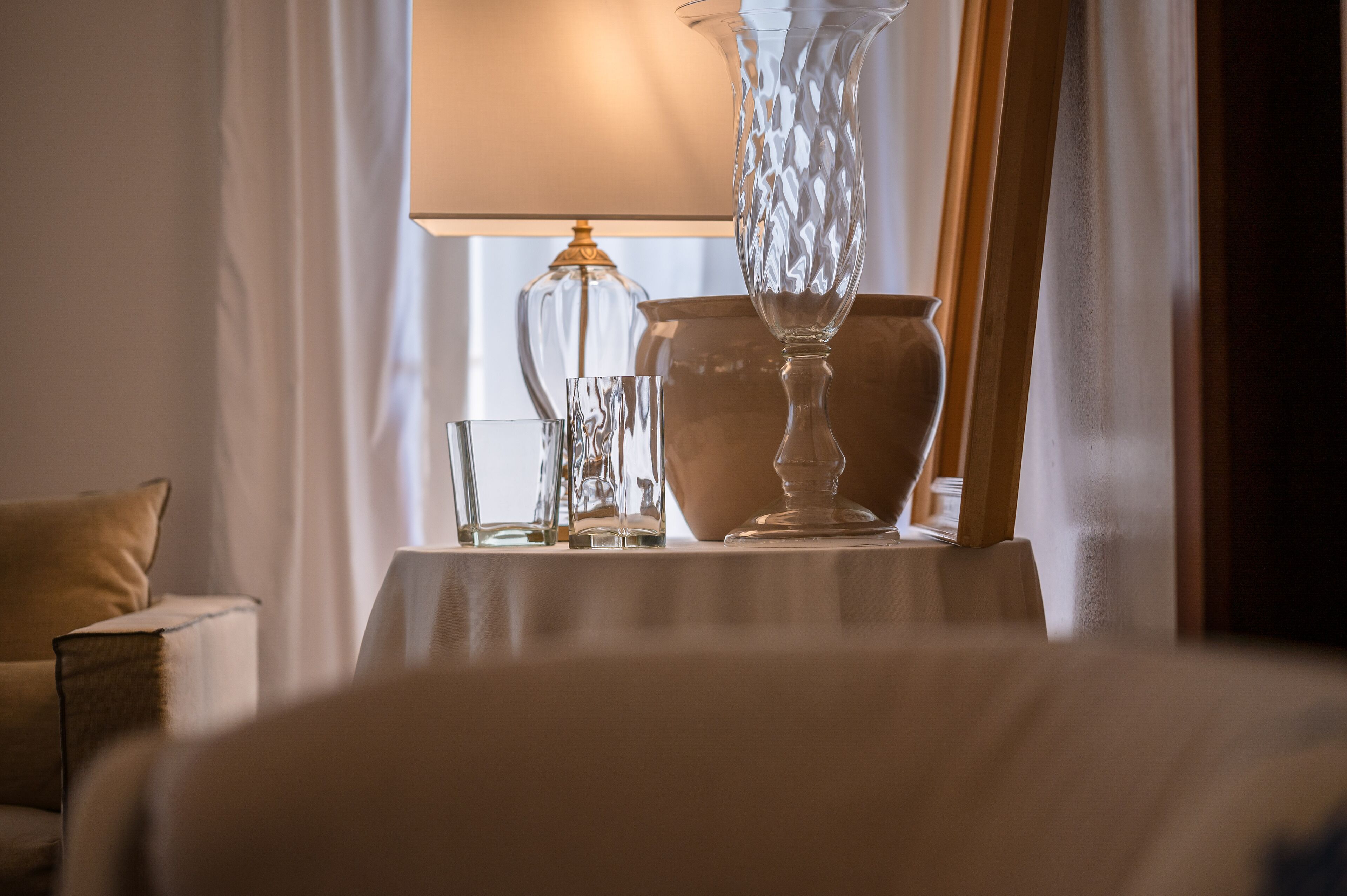 Een kamer met een gedekte tafel, helder servies, een vaas, een lamp en uitzicht op een raam met gordijnen.