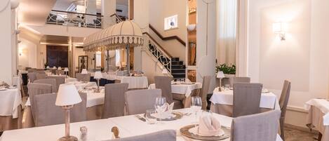 A spacious dining area with multiple tables set for guests, a grand staircase, and a chandelier.