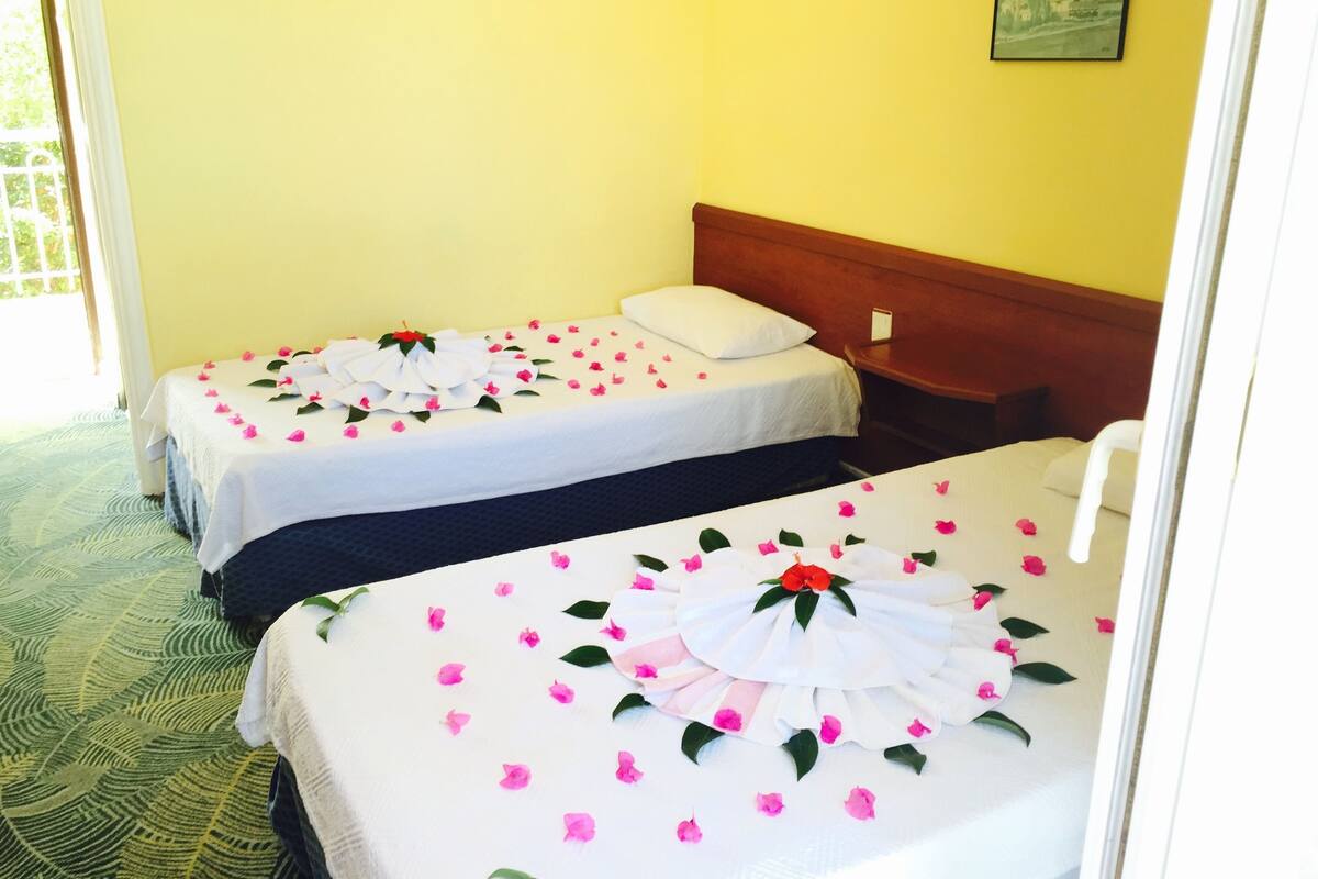 Deux lits simples avec du linge de lit blanc et des pétales de fleurs roses, une tête de lit en bois et un petit tableau encadré accroché au mur.