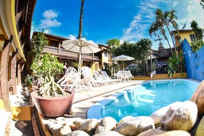 A pool area with lounge chairs, a large planter with a tree, and a blue pool. - Pousada a Flor Da Terra (Porto Seguro)