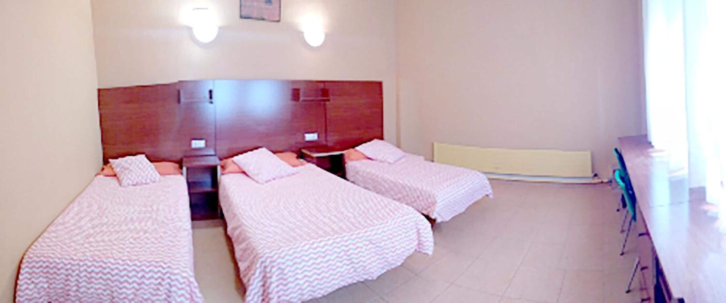 Habitación de hotel con cabecera de madera, dos camas con ropa de cama rosa, una mesita de noche y una ventana con persianas.