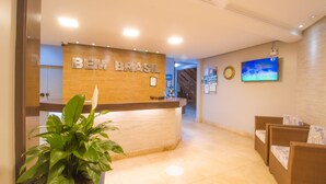 A reception area with a wooden reception desk labeled "BEM BRASIL," a potted plant, wicker chairs, and a television mounted on the wall. - Hotel Bem Brasil (Porto Seguro)