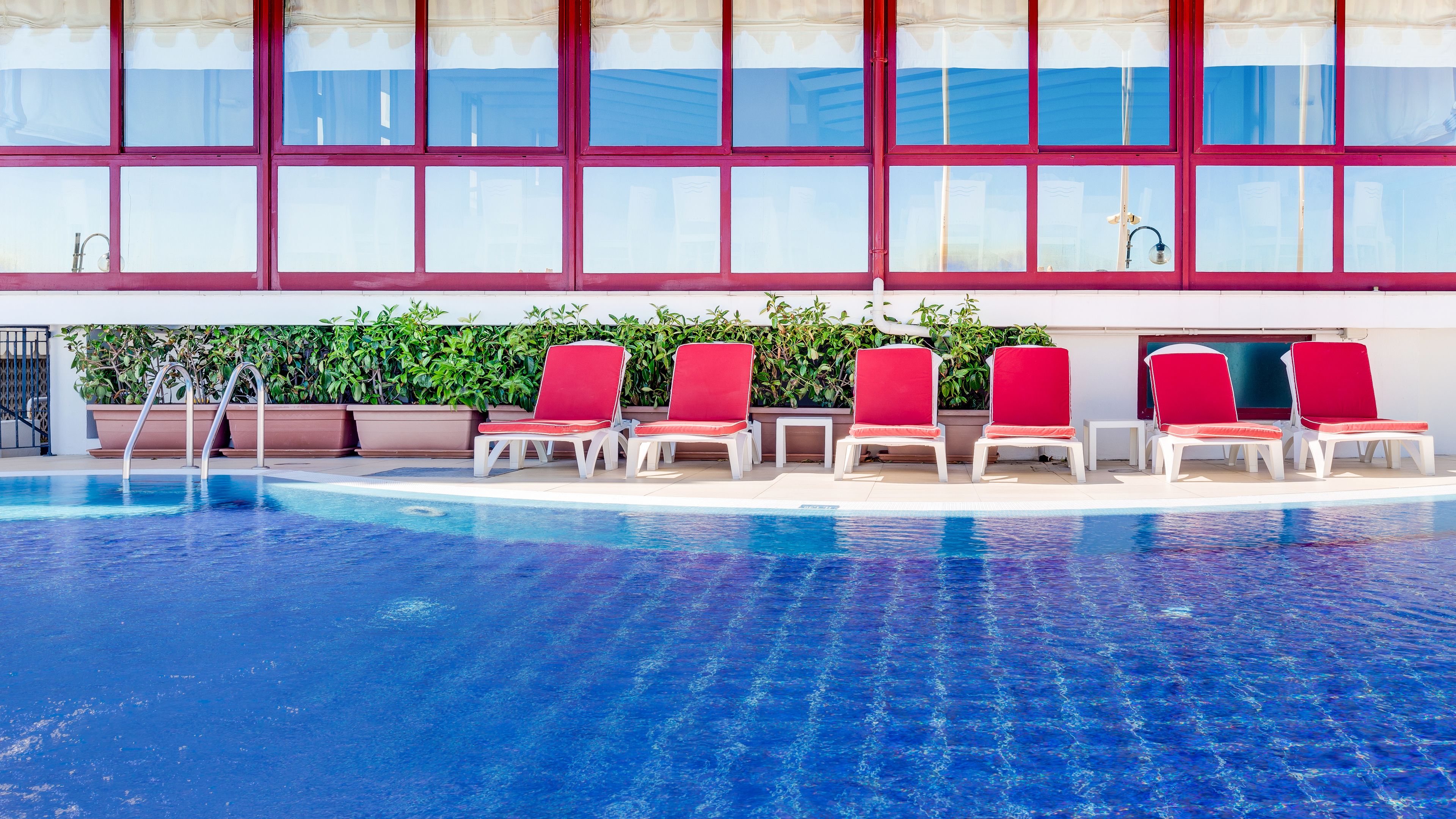 Une piscine avec des chaises longues rouges et un grand mur vitré en arrière-plan.