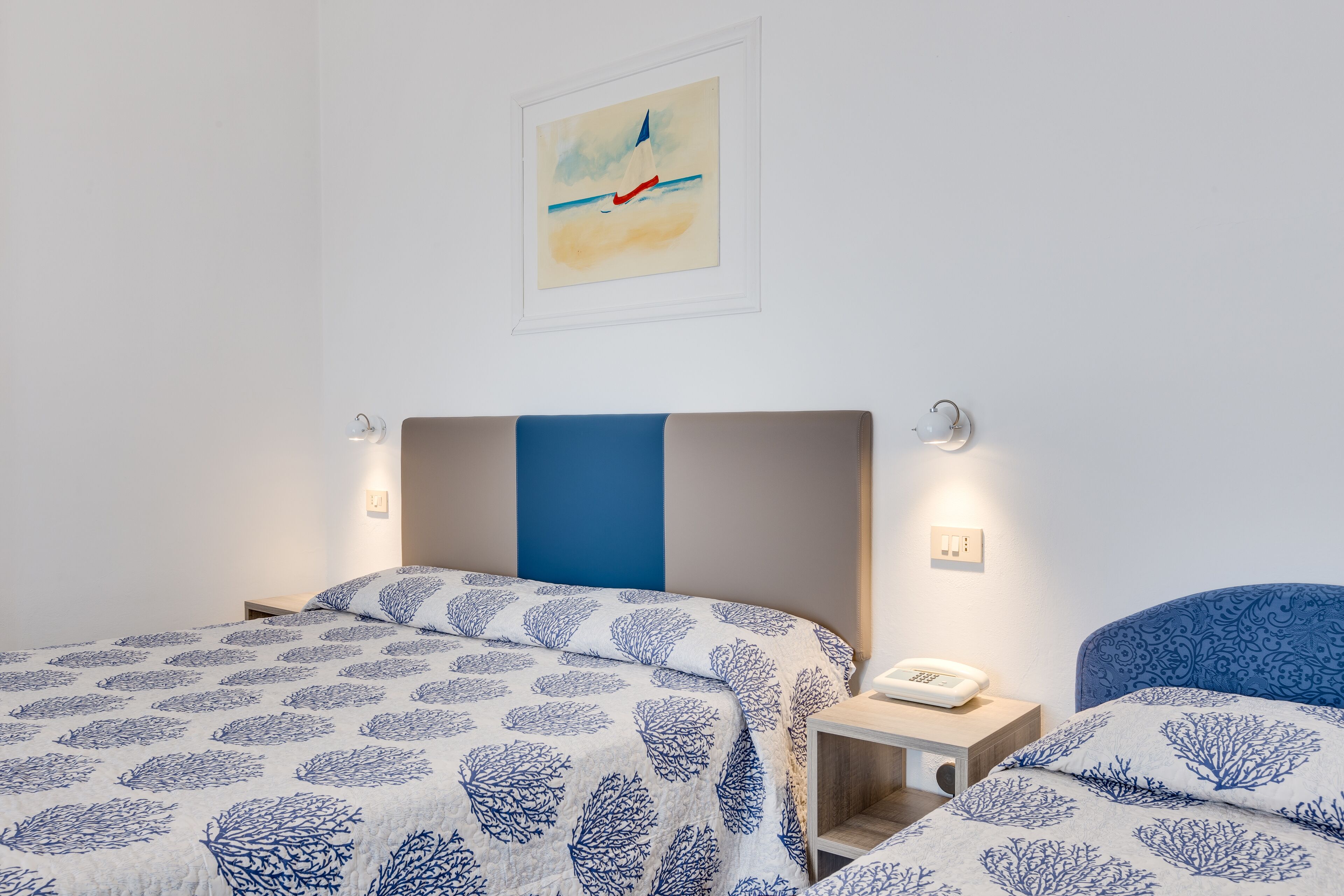 A hotel room with two beds, each with a blue and white patterned bedspread, a bedside table with a phone, and a painting of a sailboat on the wall.