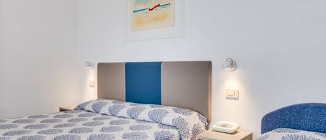 A hotel room with two beds, each with a blue and white patterned bedspread, a bedside table with a phone, and a painting of a sailboat on the wall.