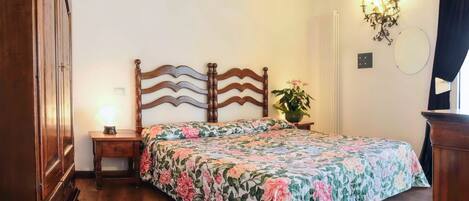 Quarto com cama de madeira, roupa de cama floral, luminária de cabeceira, planta em vaso e radiador.
