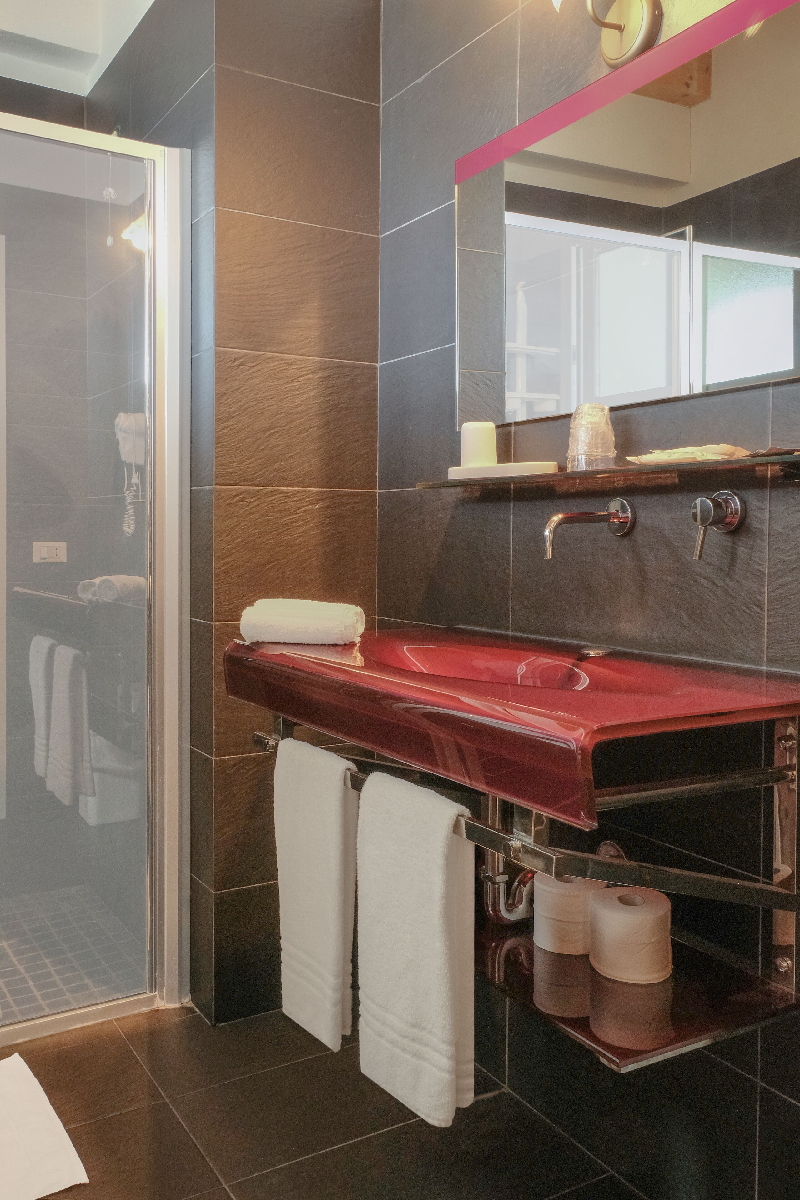 A modern bathroom with a glass shower enclosure, a red vanity, and dark tiled walls.