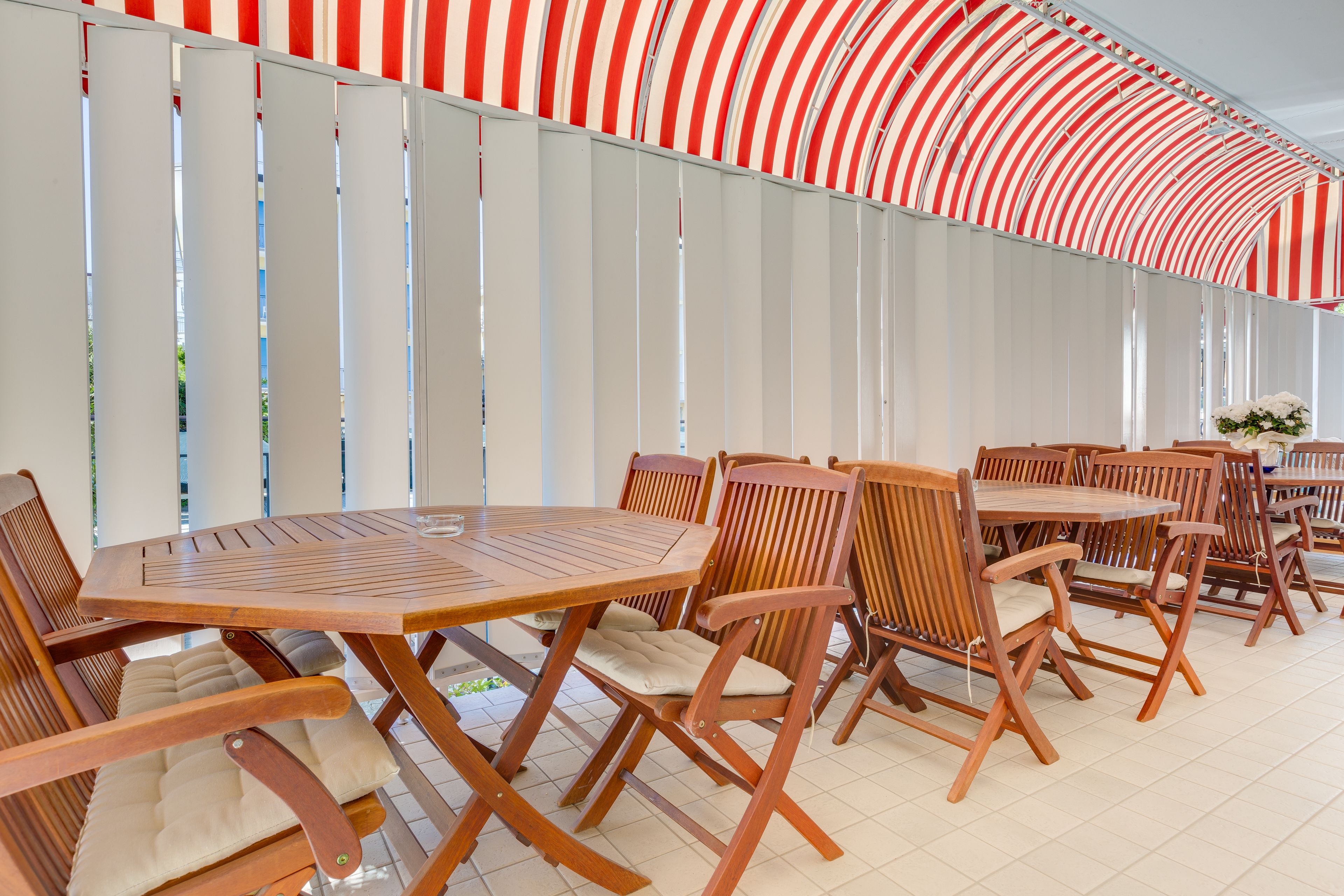 A covered outdoor seating area with wooden tables and chairs, a striped awning, and a view of the outside.