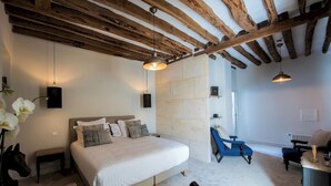 A bedroom with a large bed, wooden beams, a stone wall, a blue chair, and a desk. - Hôtel Le Chantilly (Chantilly)
