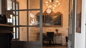 A hallway with a wooden floor, a black cabinet, and a glass door leading to a room with a stone wall, a wooden table, and a hanging light fixture. - Hôtel Le Chantilly (Chantilly)