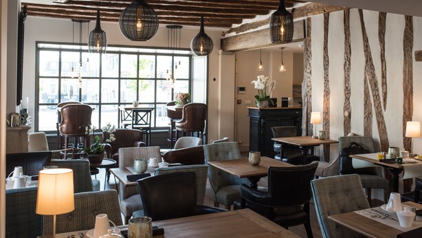 A cozy restaurant with wooden tables and chairs, hanging pendant lights, and a large window offering a view of the city. - Hôtel Le Chantilly (Chantilly)