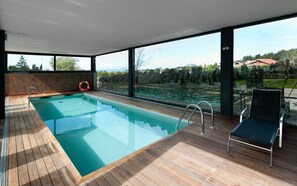 A modern indoor swimming pool with clear glass walls, wooden deck, and a lounge chair. - Mas Albereda (Sant Julia de Vilatorta)