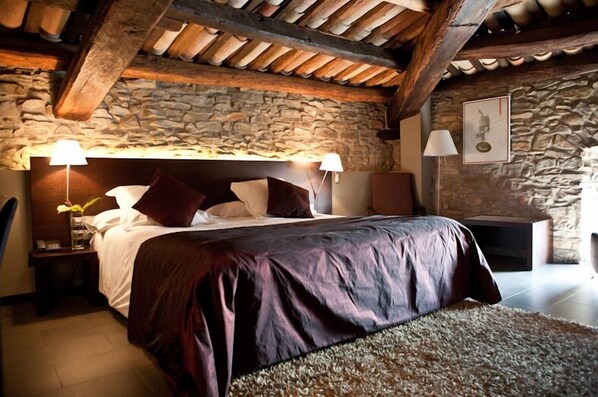 A bedroom with a stone wall, wooden beams, a large bed with a headboard, two bedside lamps, and a framed picture on the wall. - Mas Albereda (Sant Julia de Vilatorta)