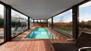 A modern outdoor swimming pool with glass walls, wooden deck, and lounge chairs.