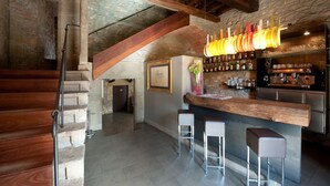 A rustic interior with exposed stone walls, wooden beams, a bar area with stools, and a staircase.
