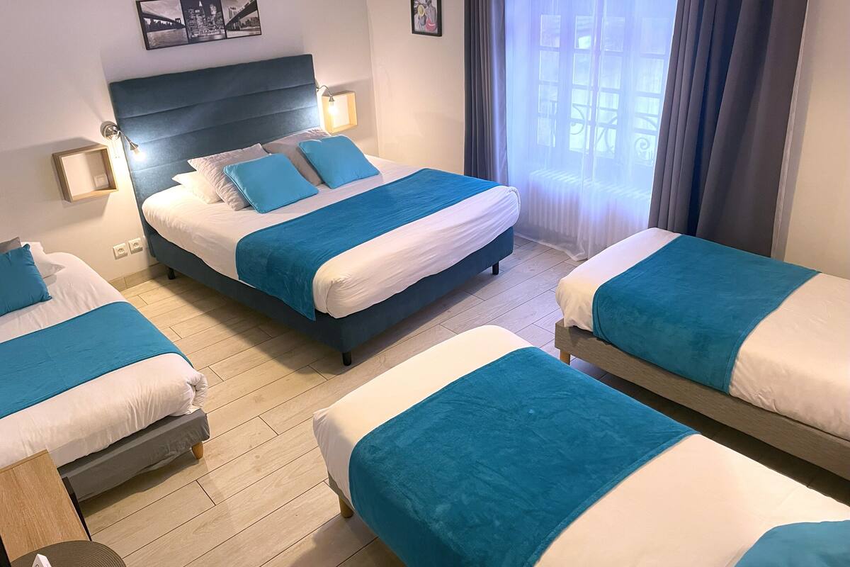 Habitación de hotel con tres camas, cada una con ropa de cama azul y blanca, piso de madera y un ventanal con cortinas.