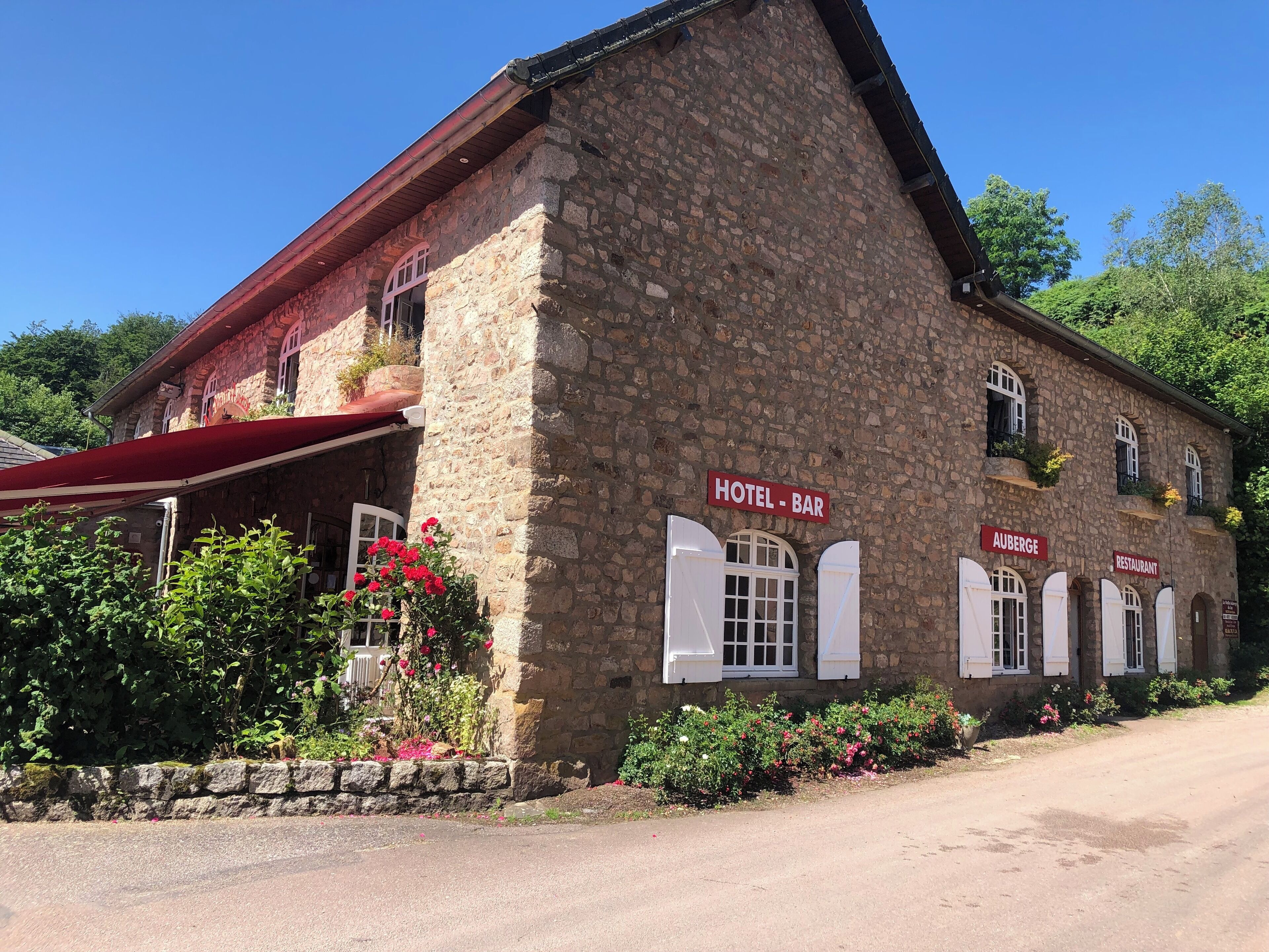 Een gebouw van steen met een rode luifel, aangeduid als hotel en bar, met witte luiken en een bord met de tekst 'Auberge Restaurant'.