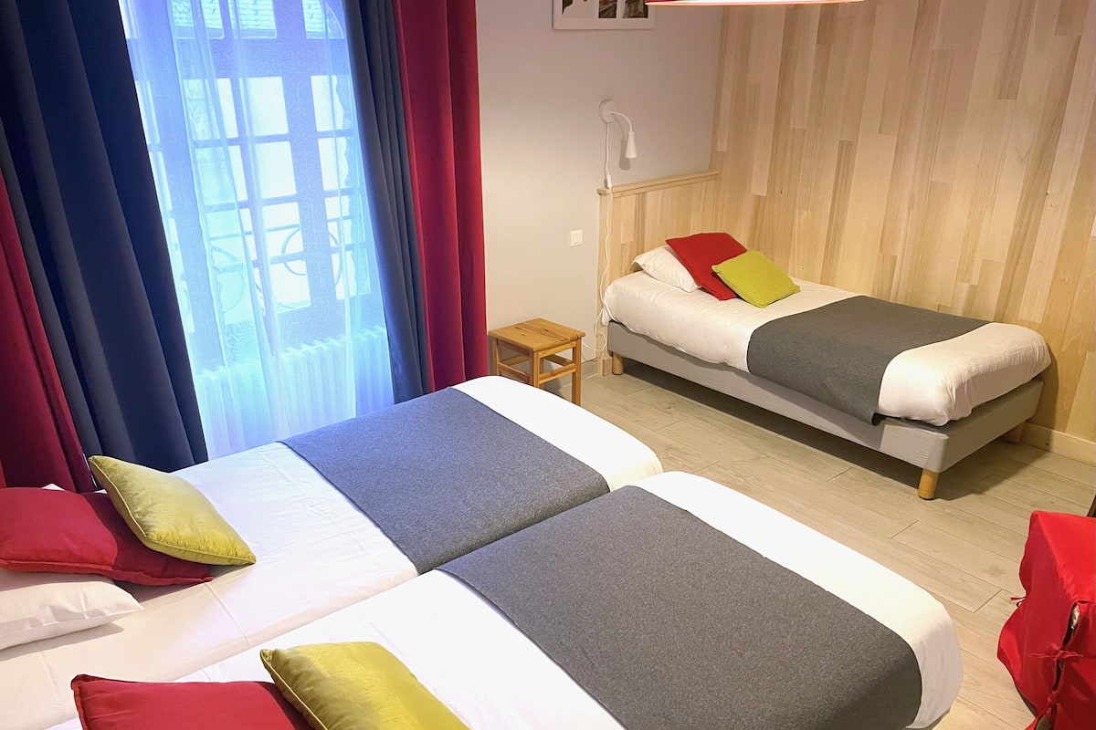 Habitación de hotel con dos camas, cada una con cubrecama gris y blanco, y almohadas rojas y amarillas. Se aprecia un pequeño tocador de madera con una lámpara, y una ventana con cortinas azules.
