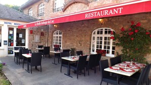 Zona de estar al aire libre de un restaurante con paredes de piedra y un toldo rojo. Las mesas están preparadas con manteles blancos y sillas negras.
