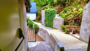 Una escalera de hormigón lleva a un pequeño edificio blanco con techo azul, rodeado de follaje y un muro de piedra.