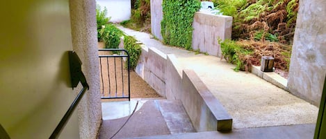 A concrete staircase leading to a small white building with a blue roof, surrounded by greenery and a stone wall.