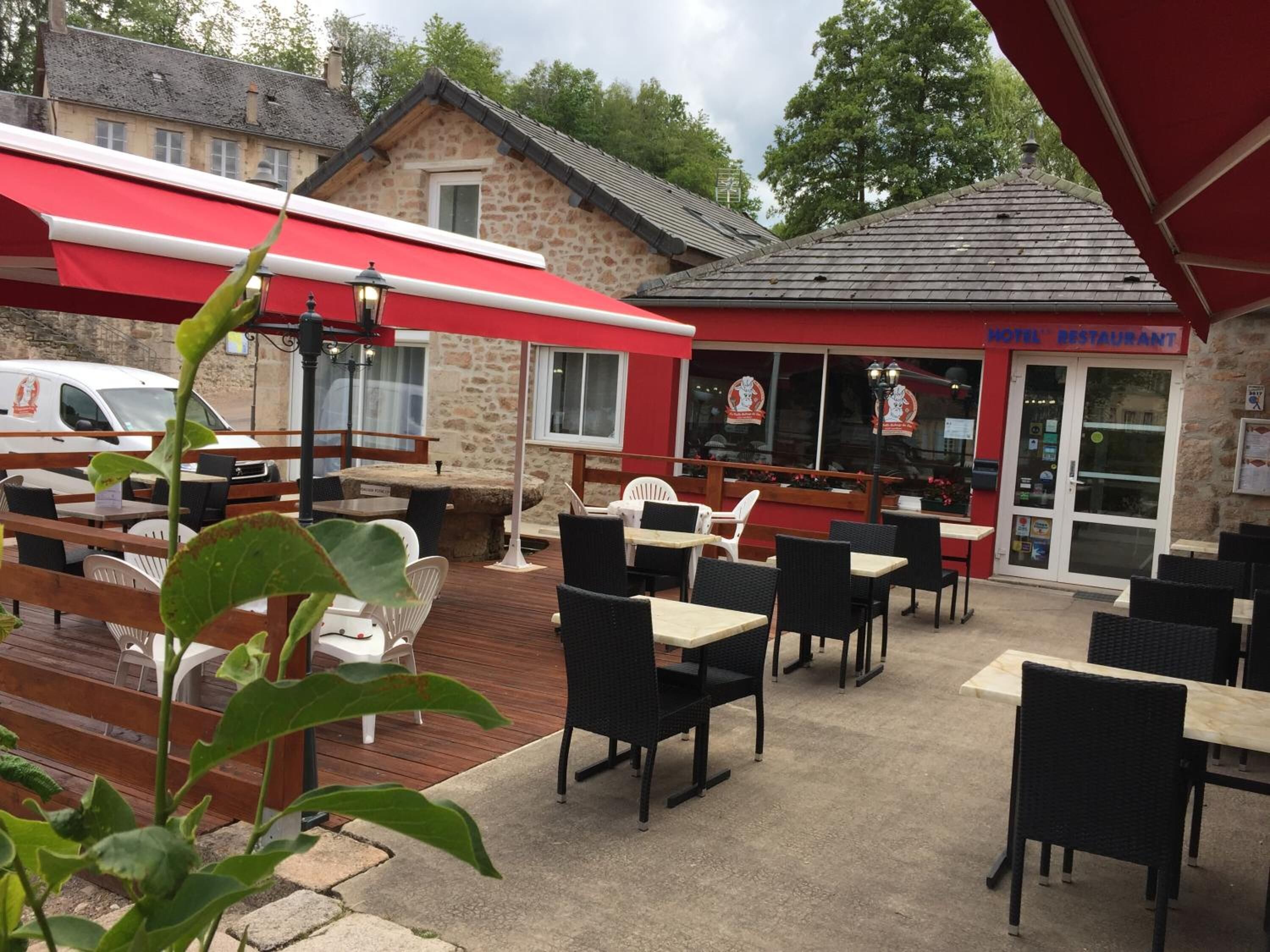Un espace extérieur aménagé avec des tables et des chaises, un bâtiment en pierre surmonté d’une marquise rouge, et une enseigne indiquant le restaurant de l’hôtel.