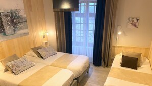 Habitación de hotel con dos camas, un ventanal con cortinas, un cuadro en la pared y una mesita con lámpara.