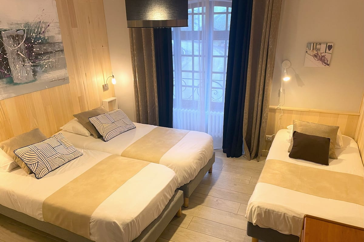 Habitación de hotel con dos camas, un ventanal con cortinas, un cuadro en la pared y una mesita con lámpara.