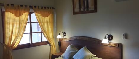 A bedroom with a wooden headboard, two bedside lamps, a bed with pillows, a window with yellow curtains, and a painting on the wall.