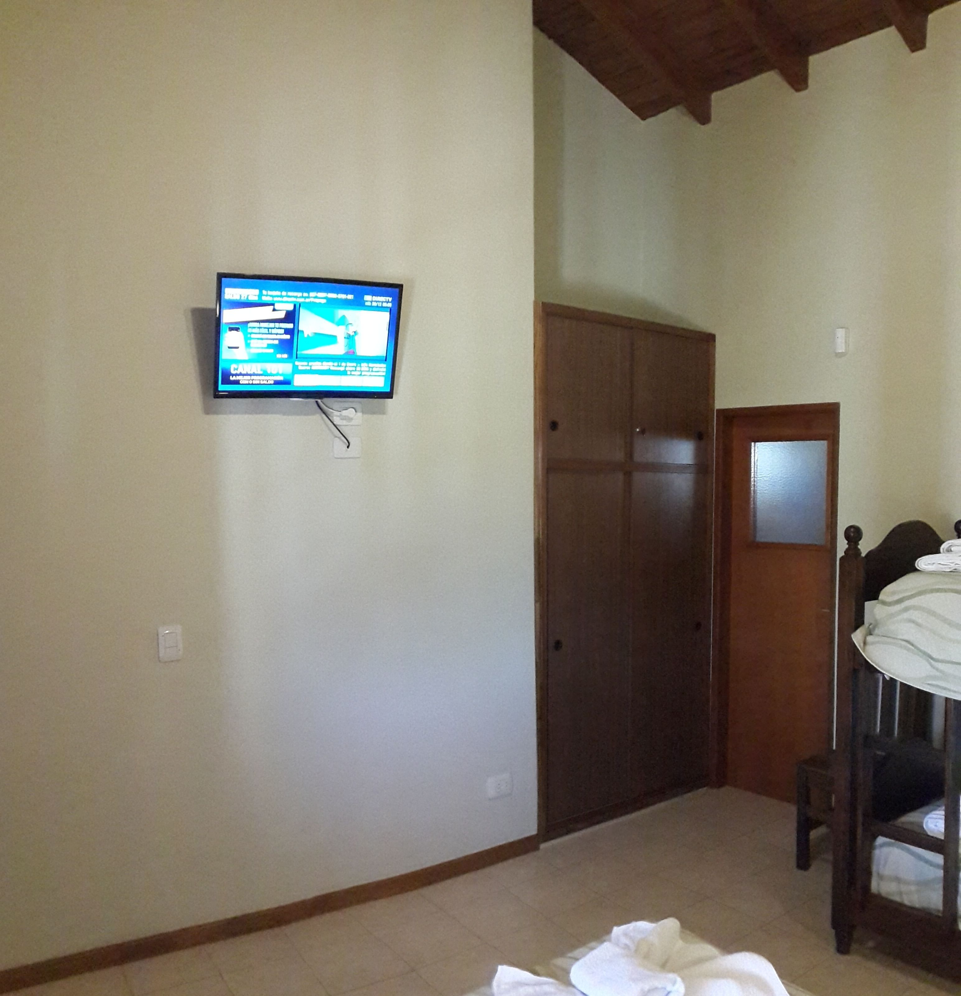 Ein Schlafzimmer mit an der Wand montiertem Fernseher, einem Holzschrank und einem Bett mit Kopfteil.