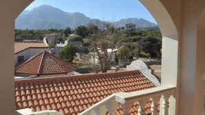 Vista a través de una ventana arqueada de un edificio con techo de tejas, barandilla blanca y montañas al fondo.
