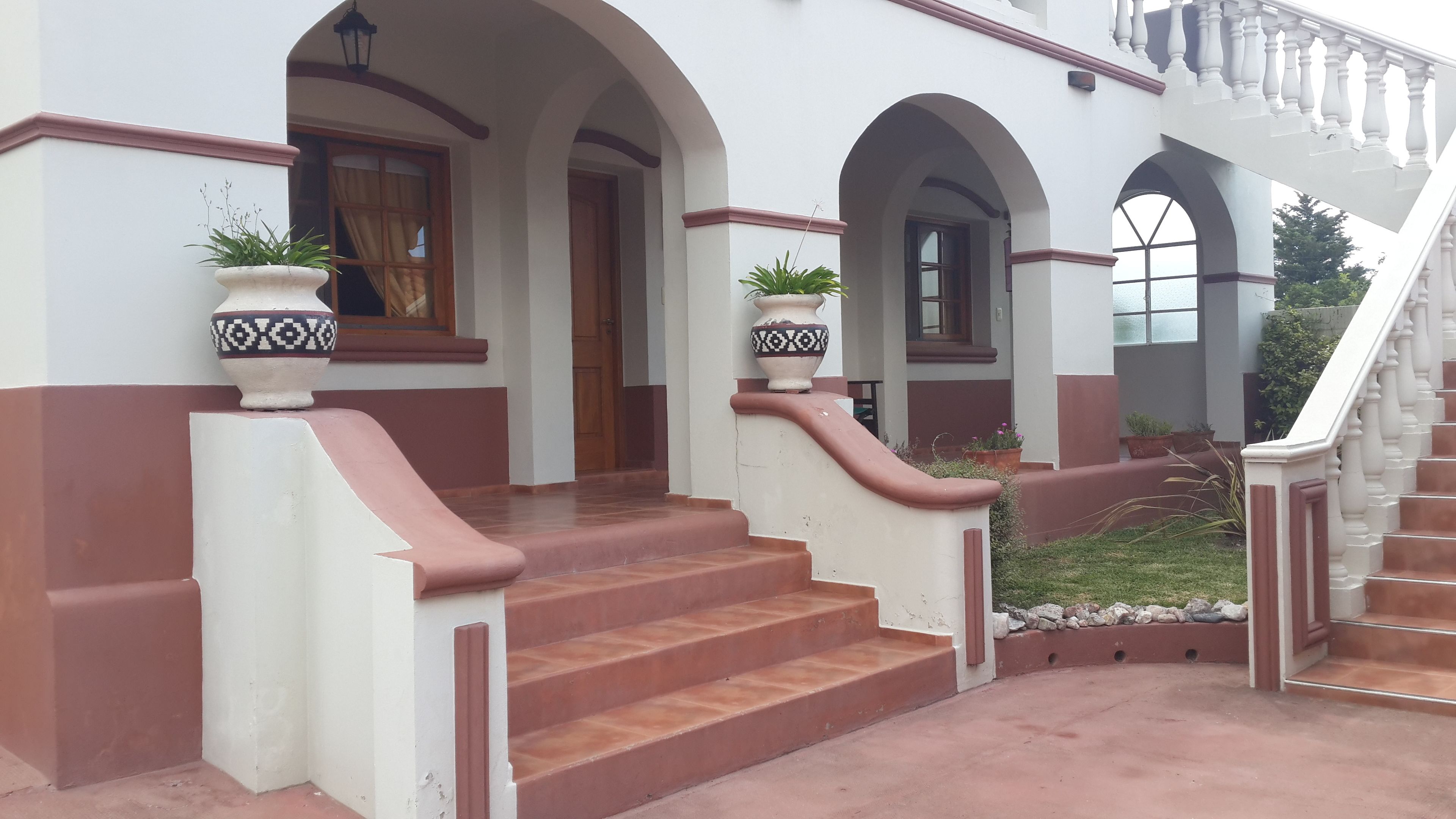 A grand entrance with a staircase leading to a covered area, featuring a white and brown color scheme, large windows, and potted plants.