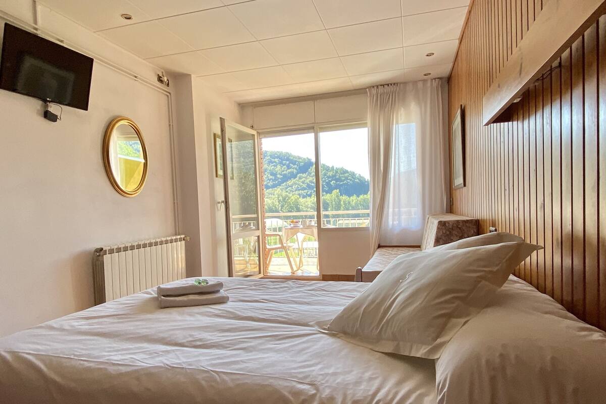 A hotel room with a large bed, a TV mounted on the wall, a mirror, and a view of a balcony with a scenic landscape.
