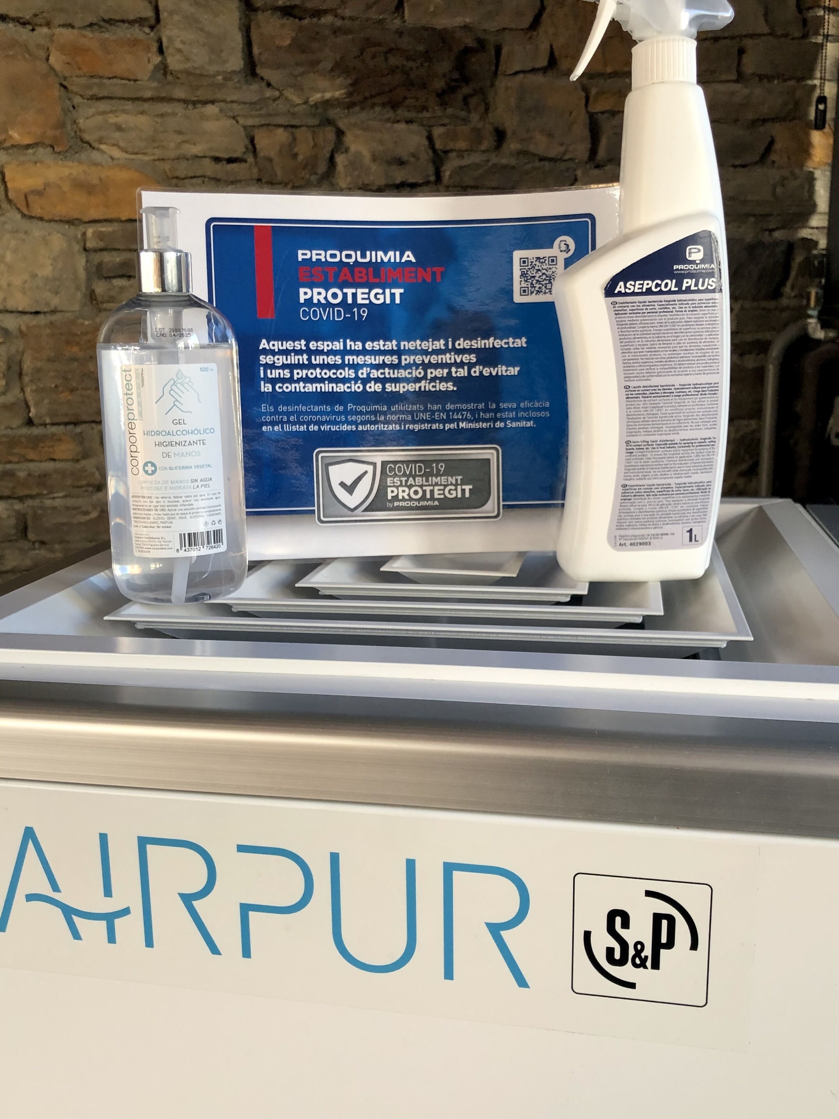Una botella de desinfectante, un atomizador y una caja con la etiqueta "PROQUIMIA ESTABLIMENT PROTEGIT COVID-19" sobre una encimera con un cartel que dice "AIRPUR" y un logo "S&P".