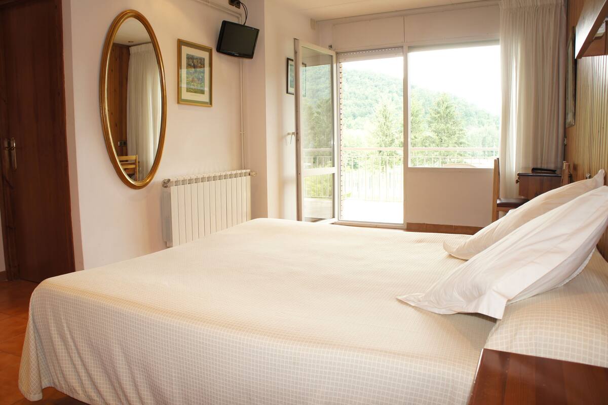 A bedroom with a large bed, a wooden door, a television on the wall, and a window with a view of trees.