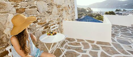 A person seated on a white chair on a stone-tiled terrace with a stone wall backdrop, a small table with fruit, and a view of a body of water and hills.