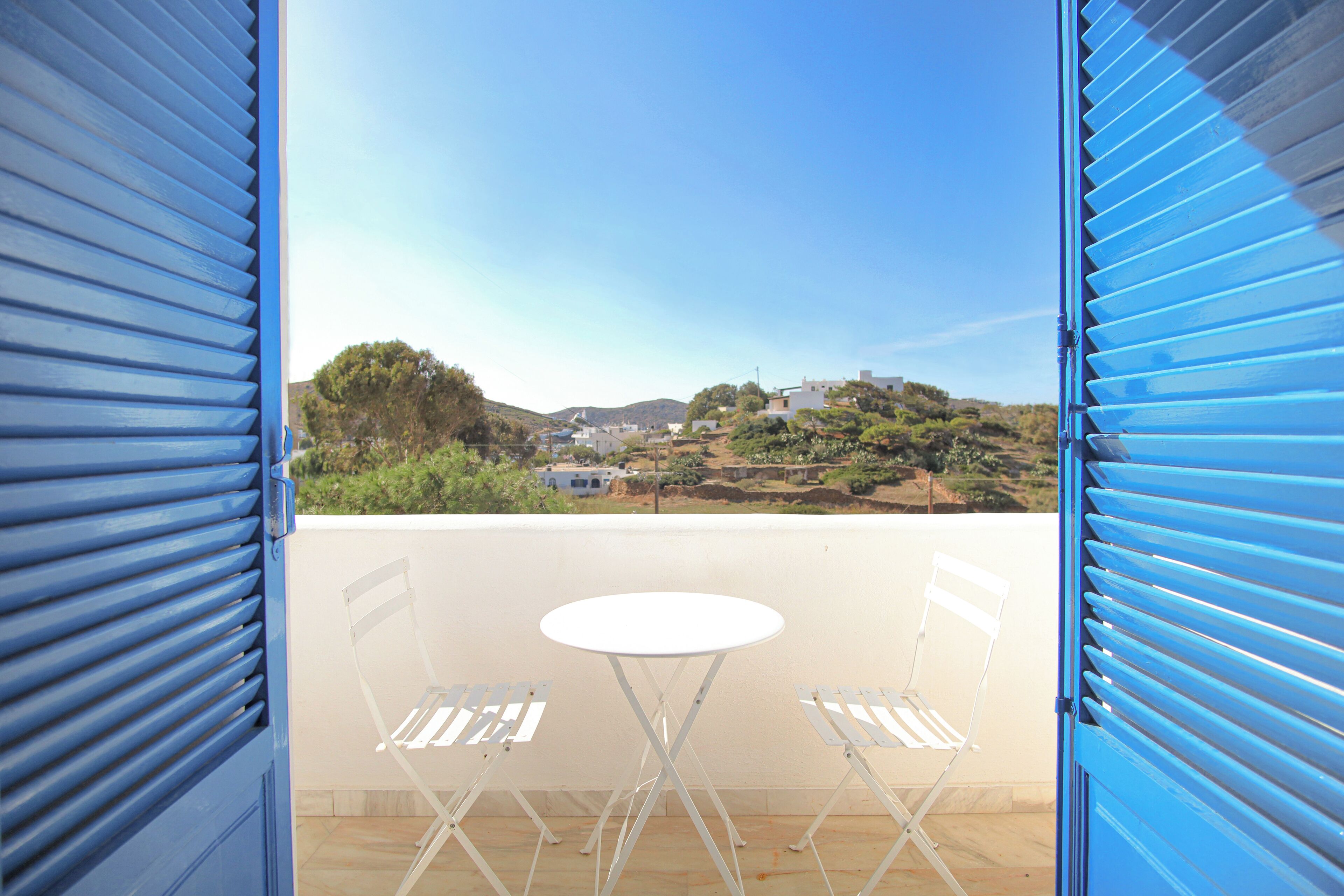 A balcony with a white table and two chairs, overlooking a hilly landscape with buildings.