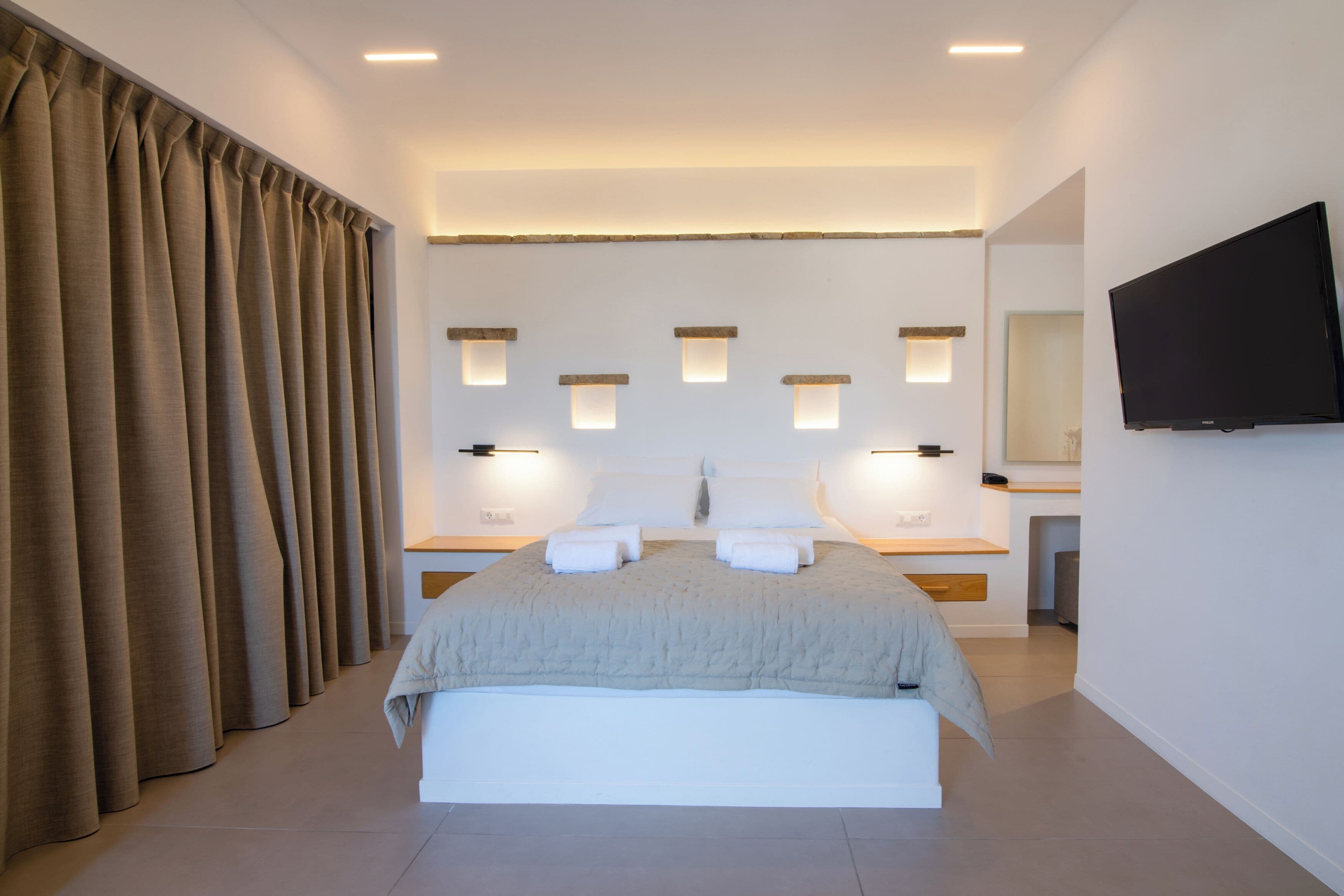 A modern bedroom with a large bed, bedside tables, a flat-screen TV, and a bathroom visible in the background.